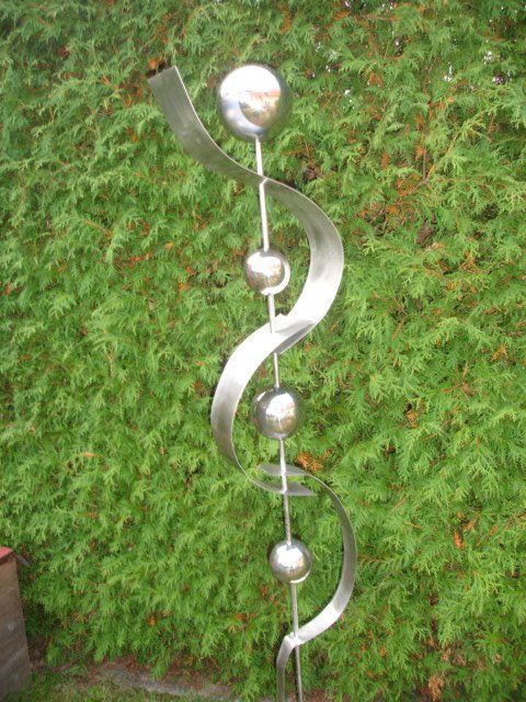 Eine Edelstahlskulptur sitzt vor einem grünen Busch.