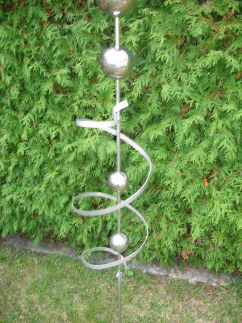 Eine Edelstahlskulptur sitzt im Gras vor einem grünen Busch.