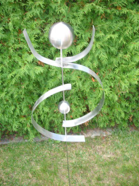 Eine Edelstahlskulptur sitzt im Gras vor einem Busch.