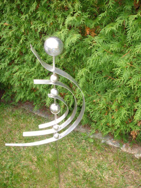 Eine Edelstahlskulptur sitzt im Gras vor einem Busch.