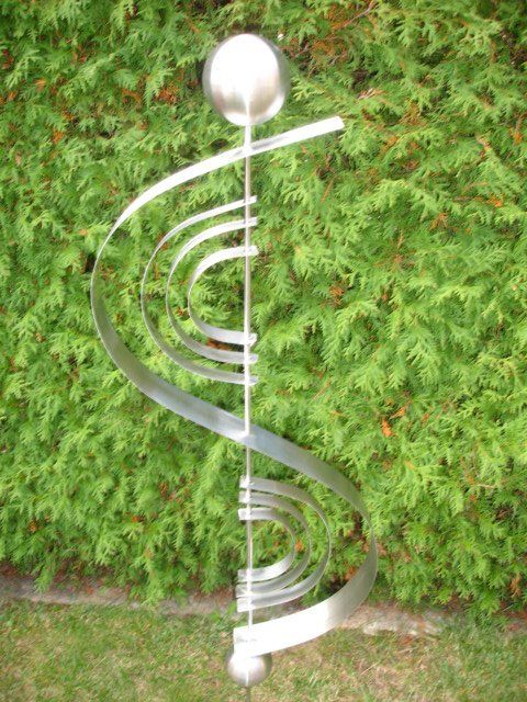 Eine Edelstahlskulptur sitzt im Gras vor einem Busch.