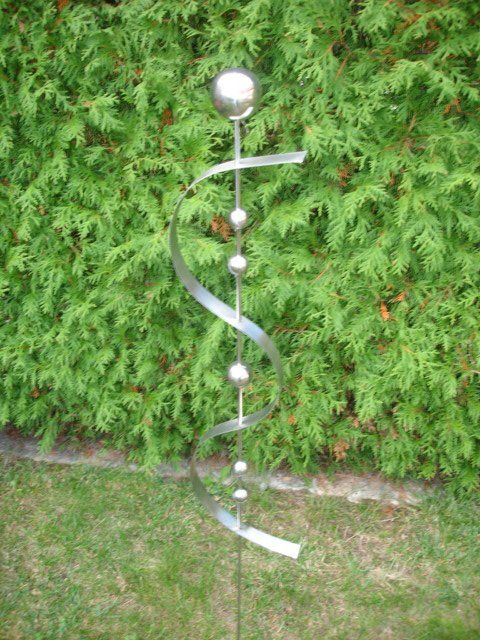 Eine Edelstahlskulptur sitzt im Gras vor einem Busch.