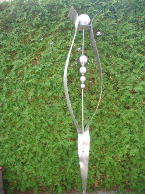 Eine Skulptur aus Edelstahl steht vor einer grünen Hecke.