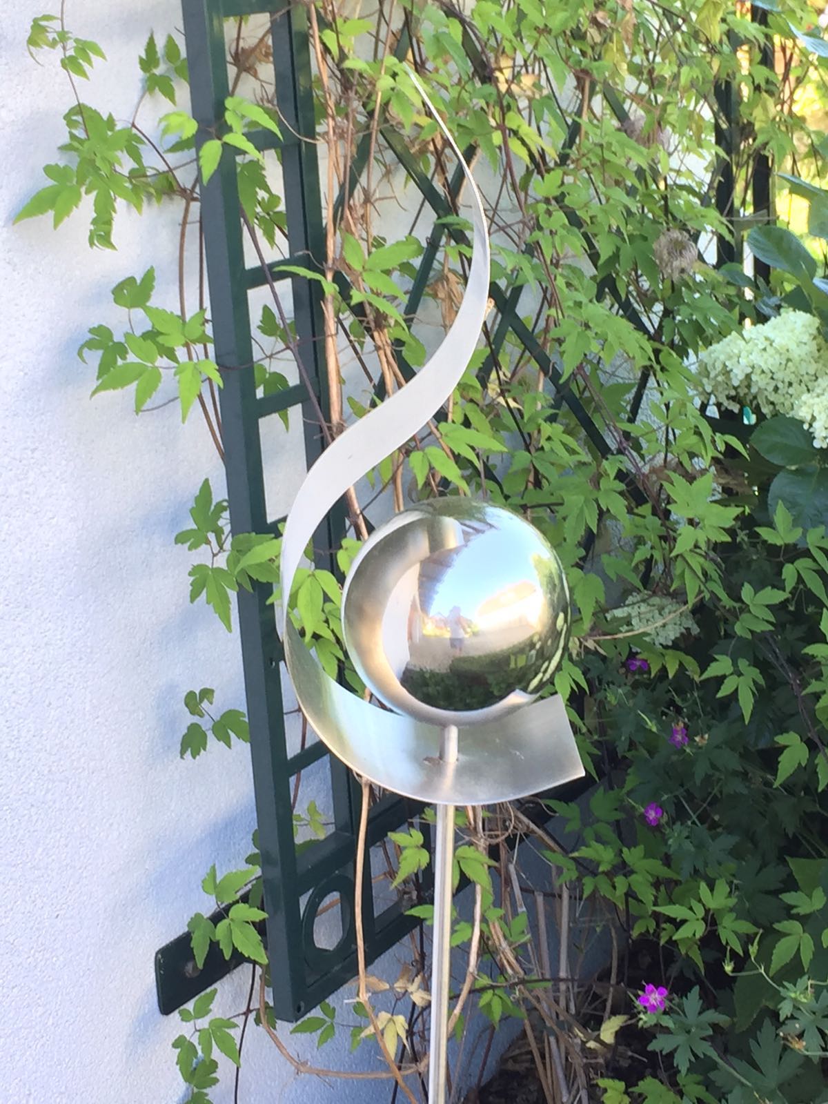 Eine Skulptur aus Edelstahl sitzt vor einem grünen Spalier.