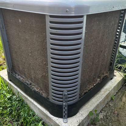 A square, gray evaporative cooler with brown filter pads sits on a concrete base outdoors.