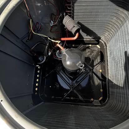 Inside view of an air conditioner unit, showing the compressor, wiring, and copper piping within a black-painted compartment.