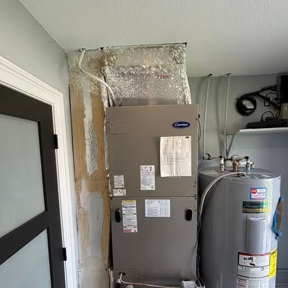 HVAC system, water heater in utility room.  Exposed ductwork, gray units.  White wall, black-framed door.