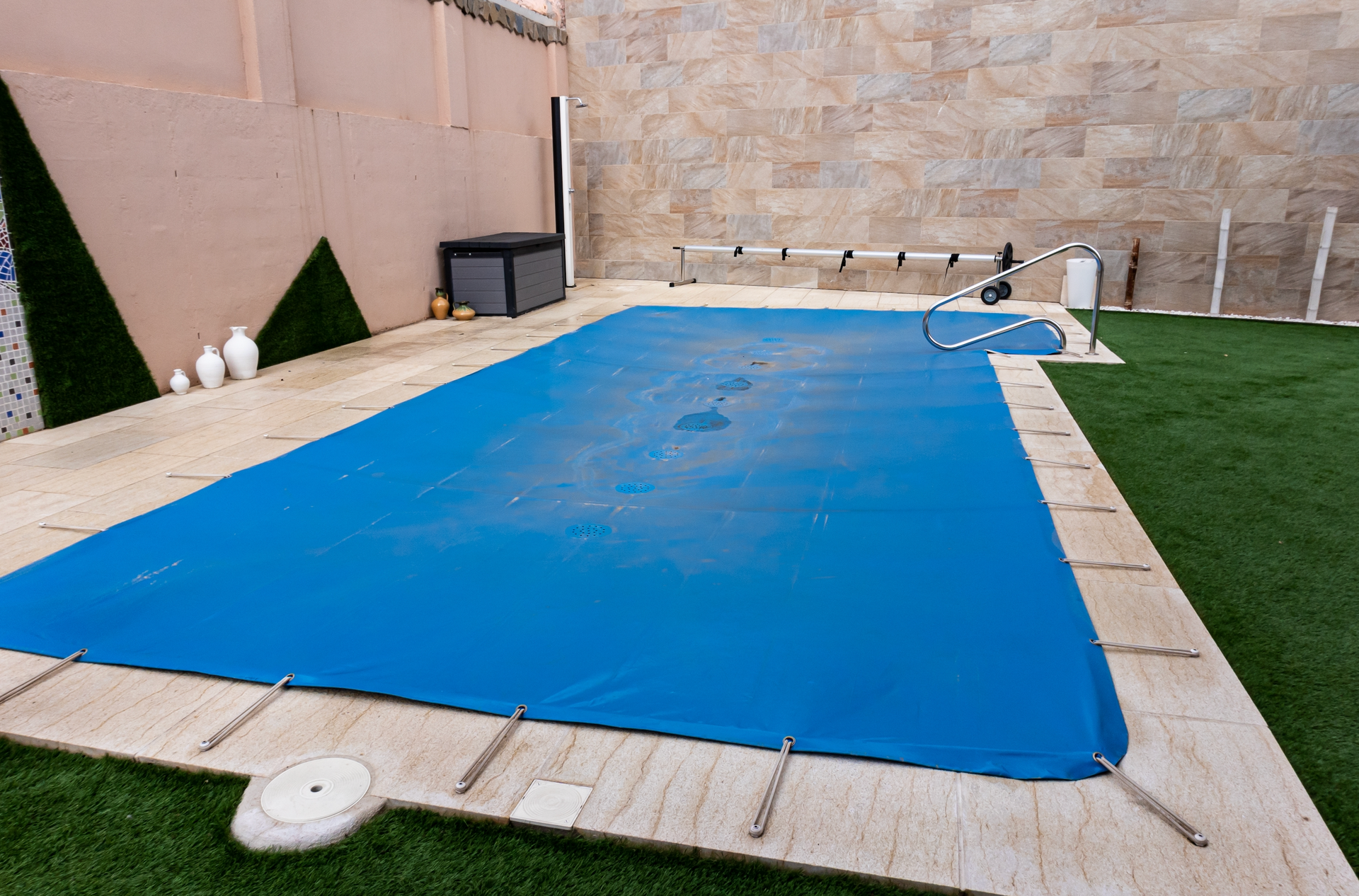Blue pool cover on a rectangular pool surrounded by beige stone and green grass.