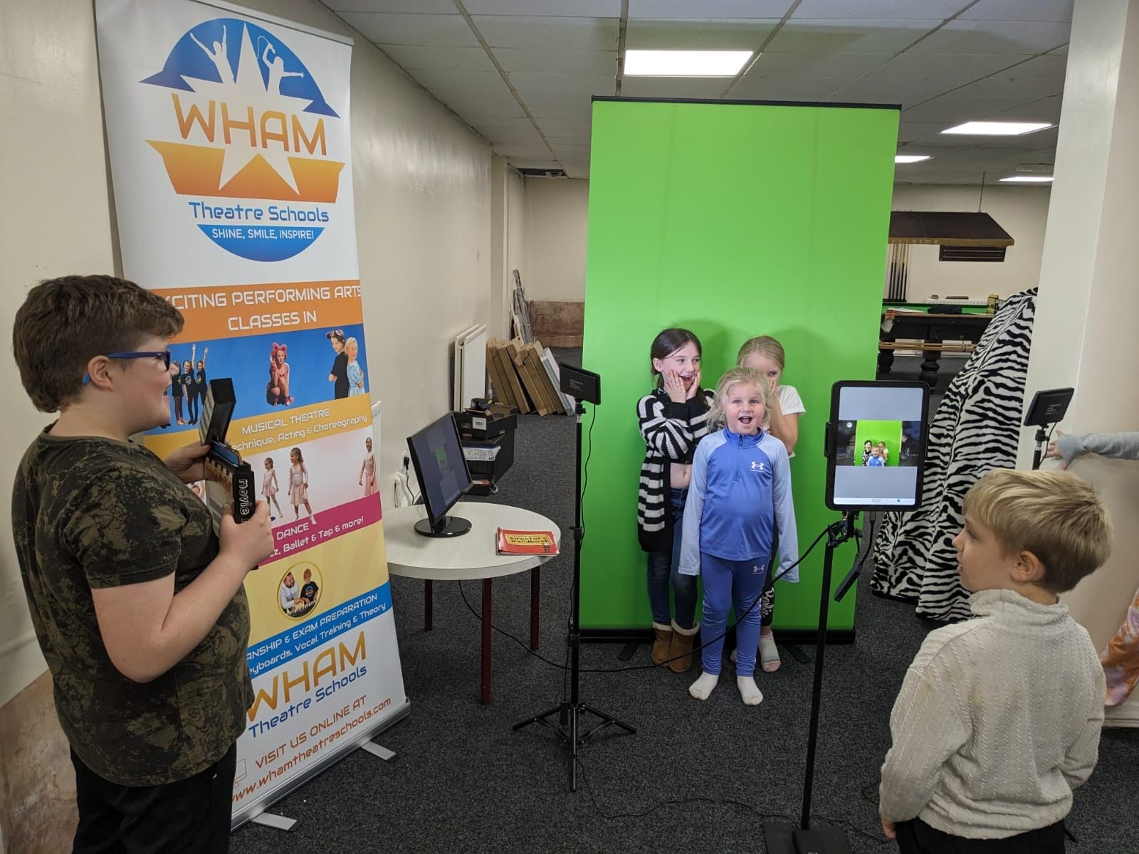 A group of children are standing in front of a green screen in front of a wham sign.