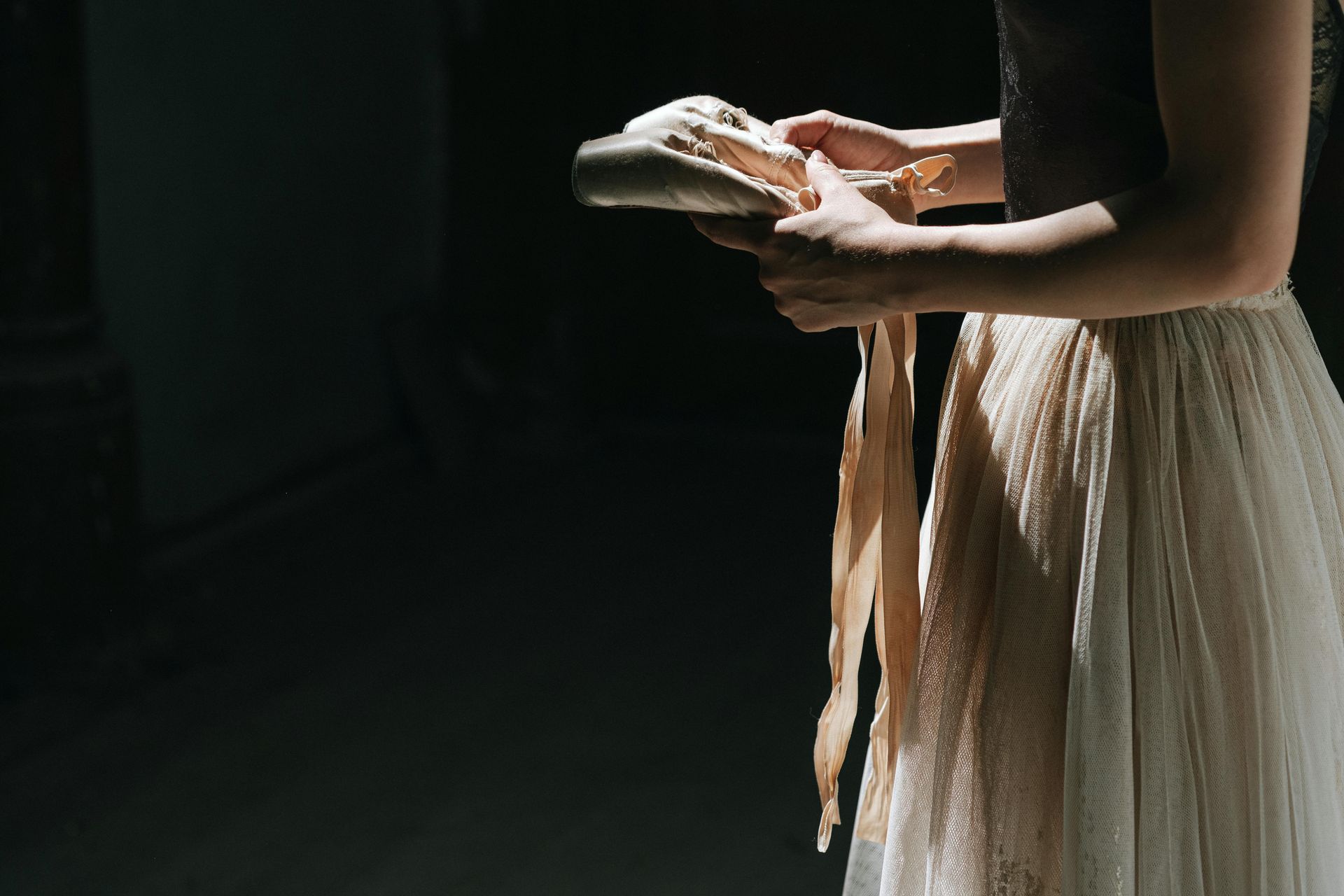 A woman in a white dress is holding a pair of pointe shoes.