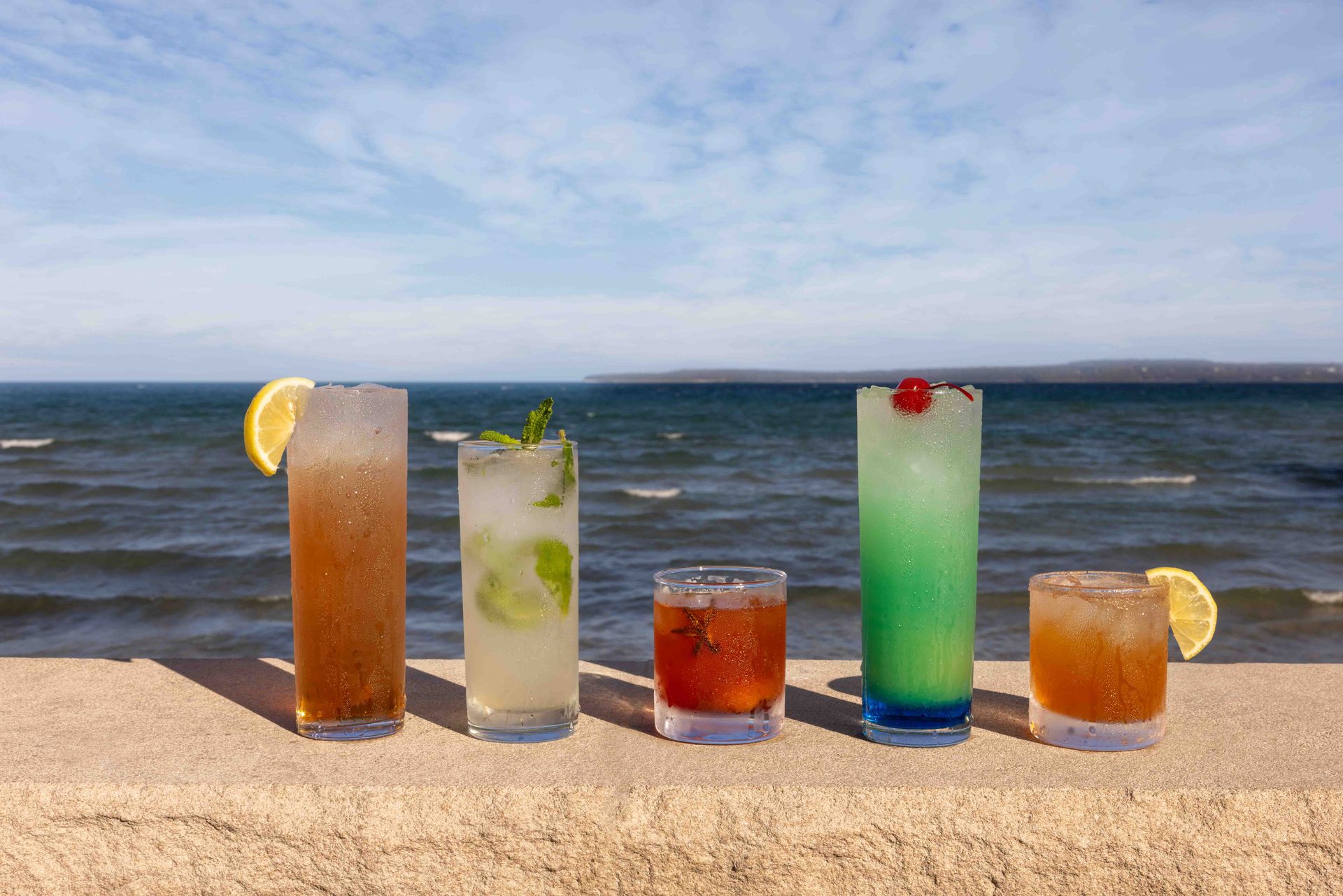 Four drinks are lined up on the beach in front of the ocean.