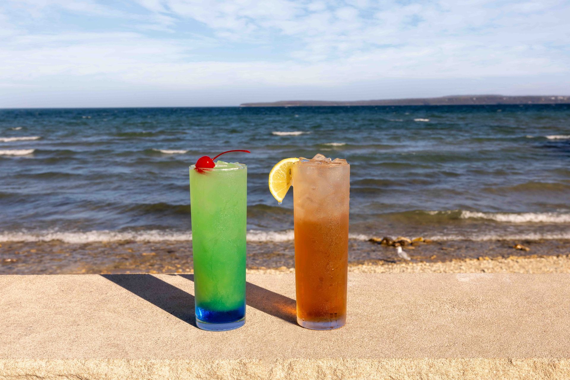 Two drinks are sitting on the beach in front of the ocean.