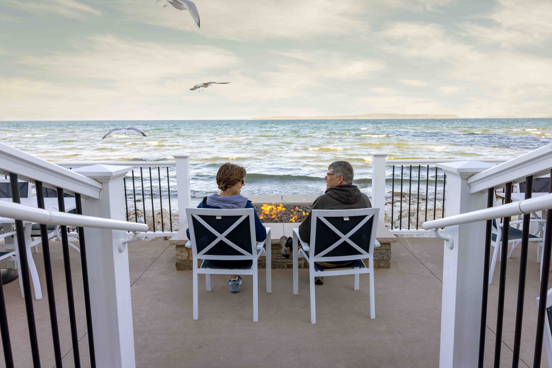 A man and a boy are sitting at a fire pit on a deck overlooking the ocean.