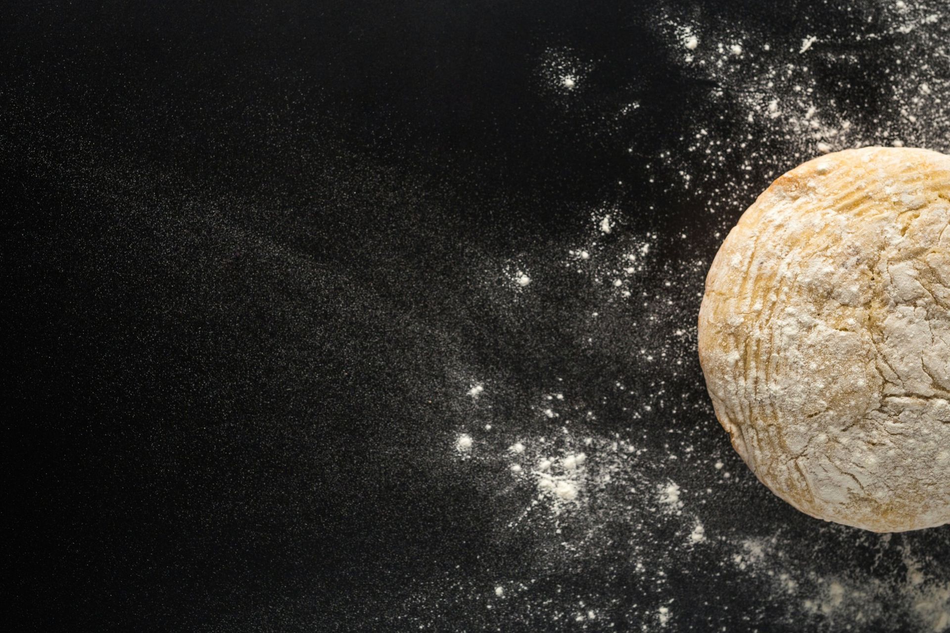 A ball of dough is surrounded by flour on a black surface.