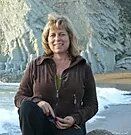 A woman in a brown jacket is sitting on a rock on the beach.