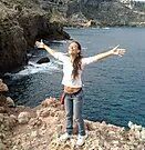 A woman is standing on a rocky cliff overlooking the ocean with her arms outstretched.