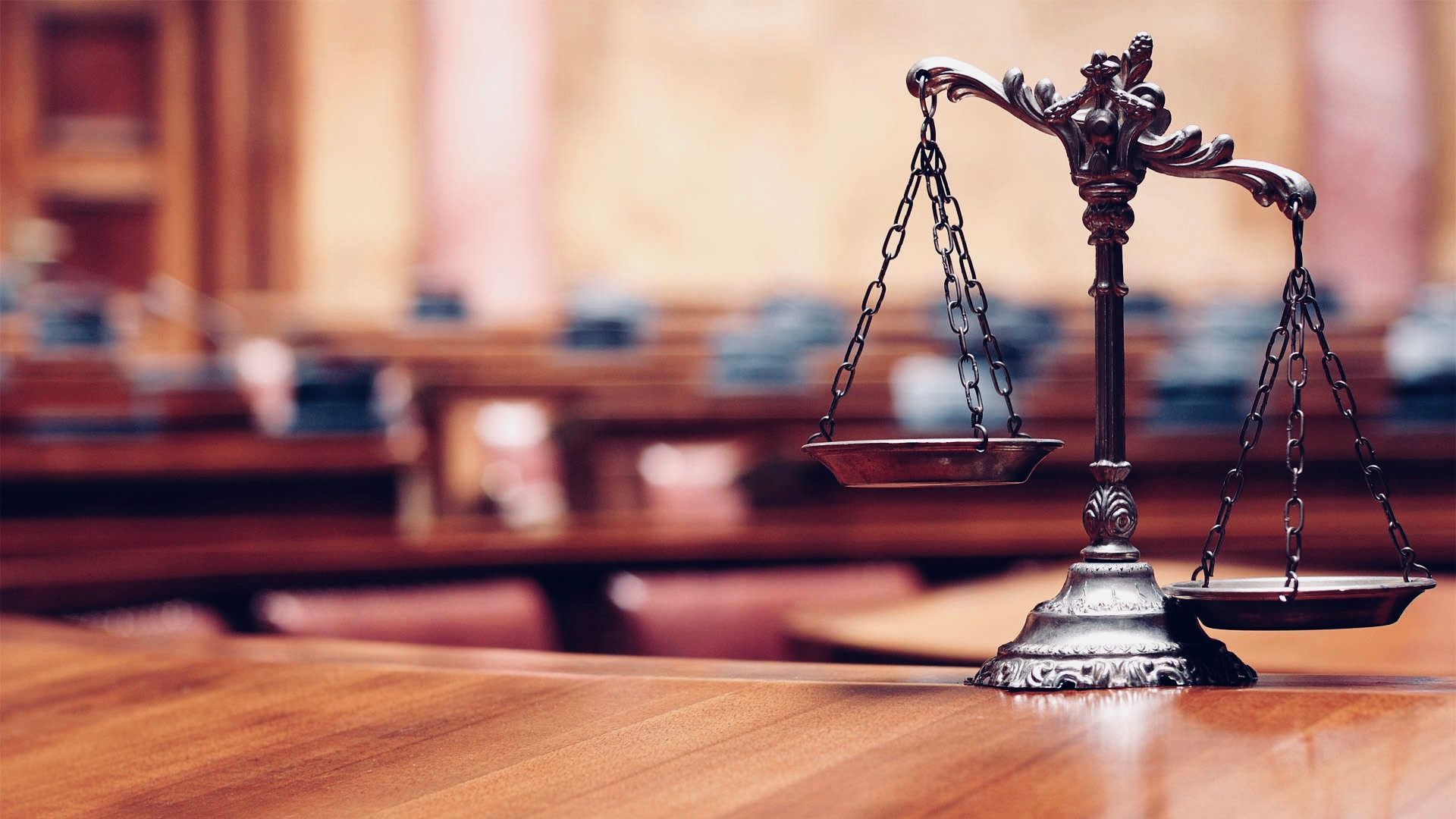 A pair of brass scales of justice sitting on a polished wooden table in a blurred courtroom setting.