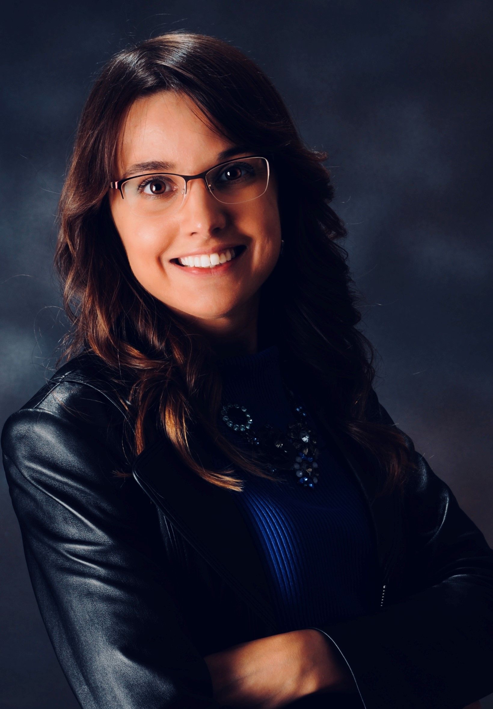 Woman with glasses smiles, arms crossed, wearing a black leather jacket and blue top.