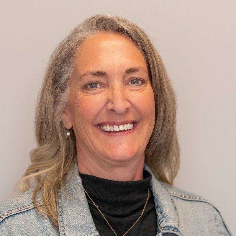 Woman with gray hair smiles, wearing denim jacket and black top, against a neutral background.