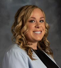 Woman with blonde curly hair, smiling, wearing a light blue jacket and black top.