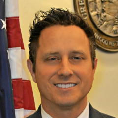 Man in a suit smiles, standing near the American flag and a gold seal.
