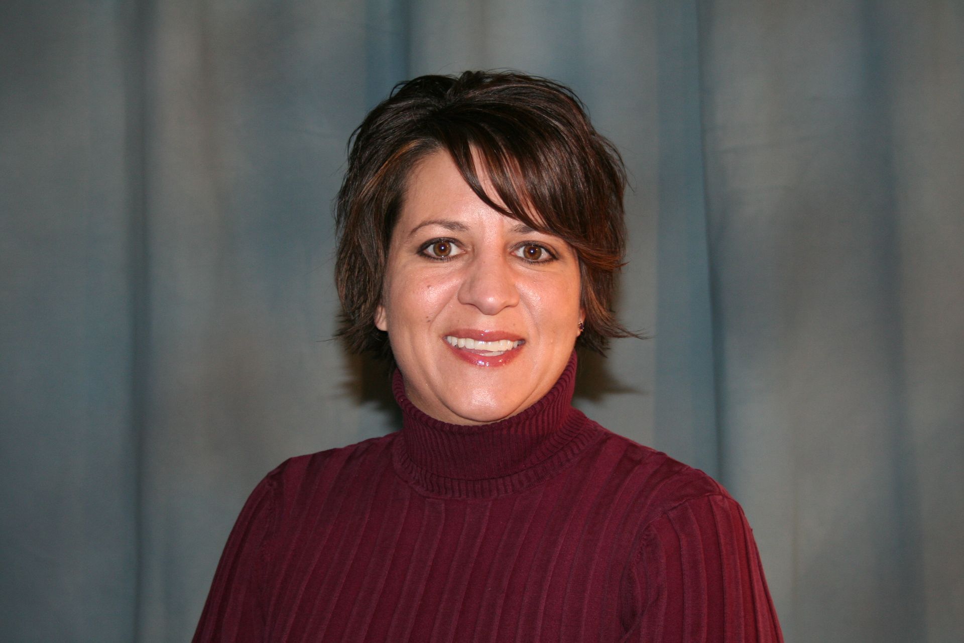 Woman in burgundy turtleneck smiles in front of blue curtains.
