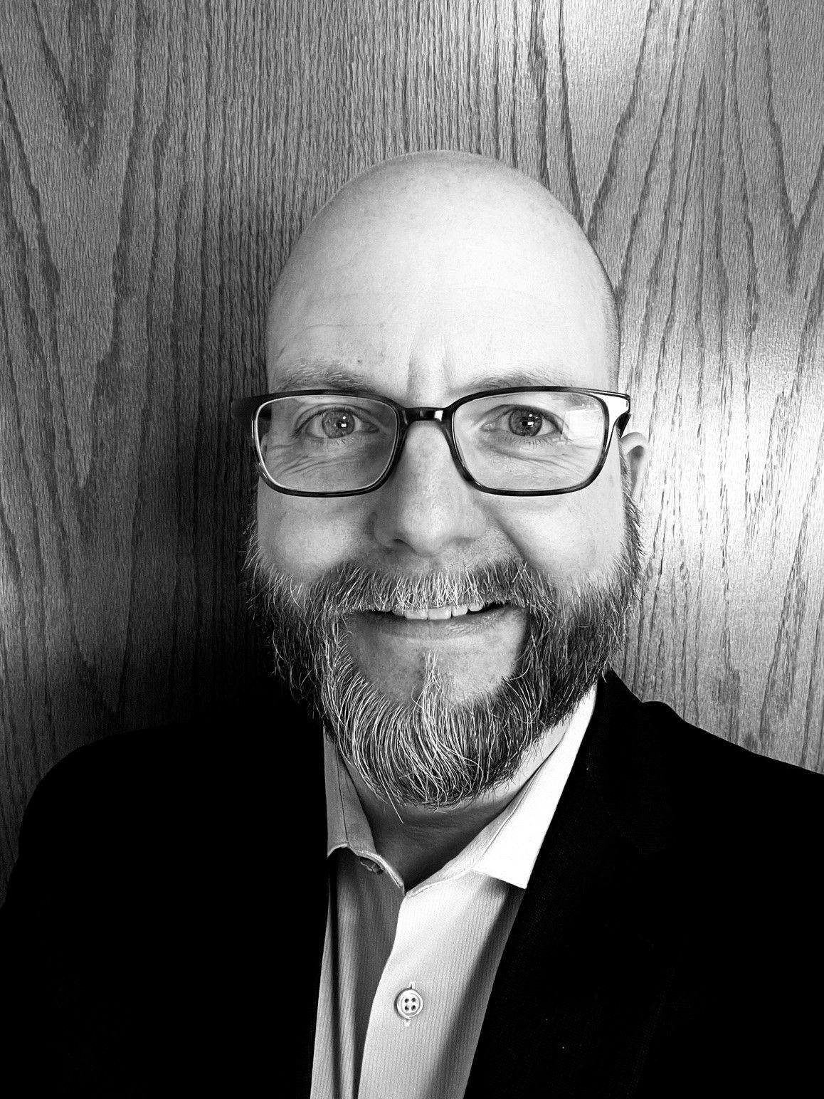Bald man with glasses and beard, smiling. Wearing a black suit jacket and collared shirt, against a wood-paneled wall.