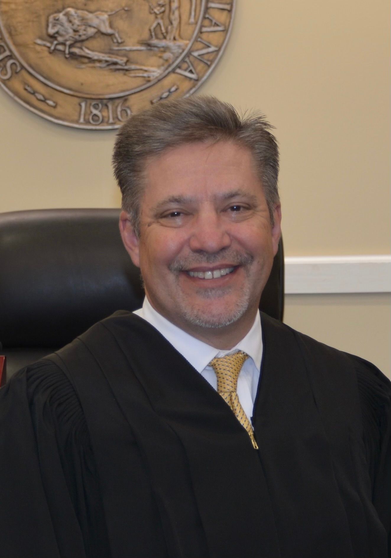 A smiling judge in a black robe and yellow tie, seated in front of an official Indiana state seal.