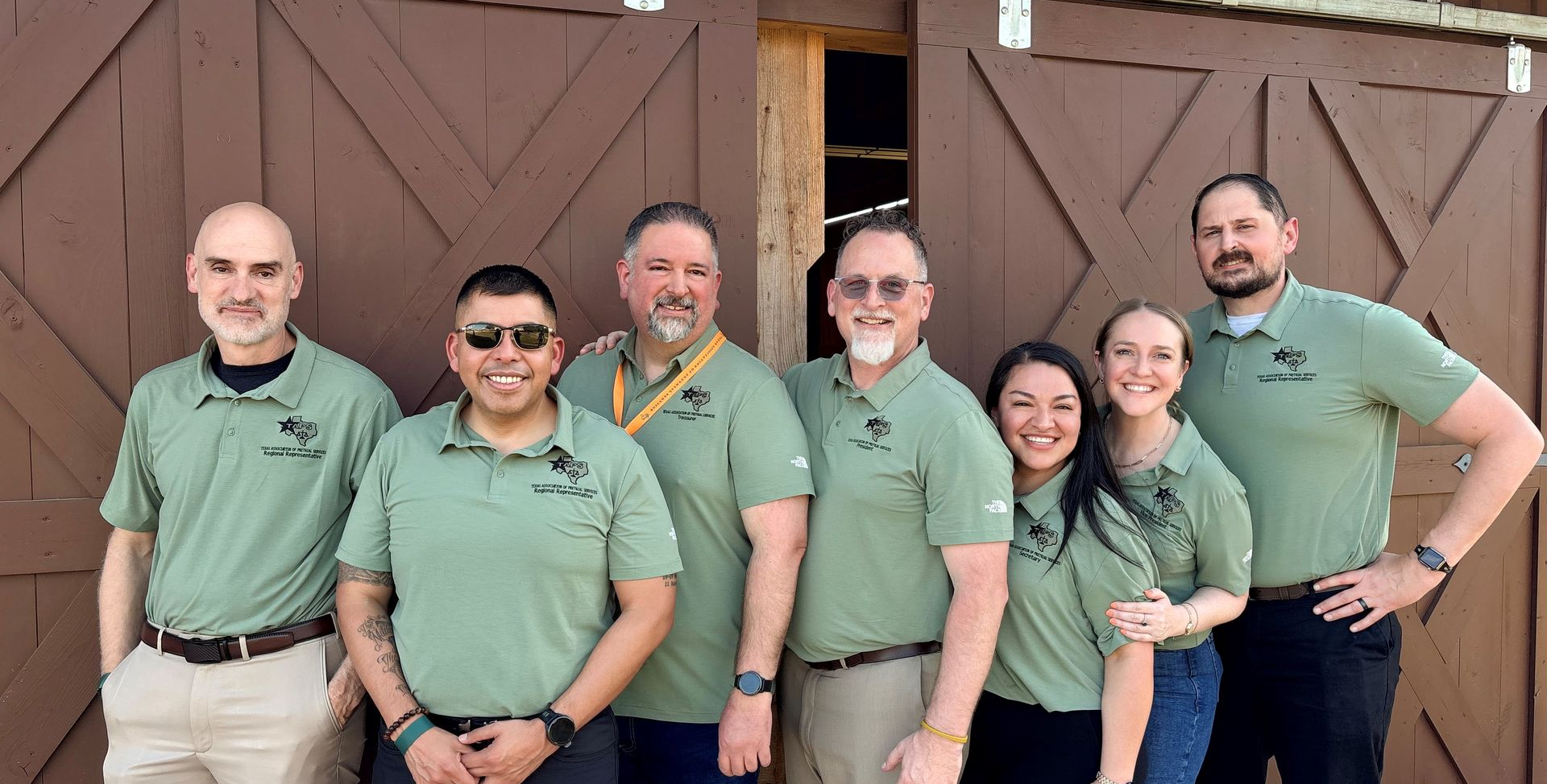 Seven people in matching green polo shirts stand in a row smiling in front of brown wooden double doors.