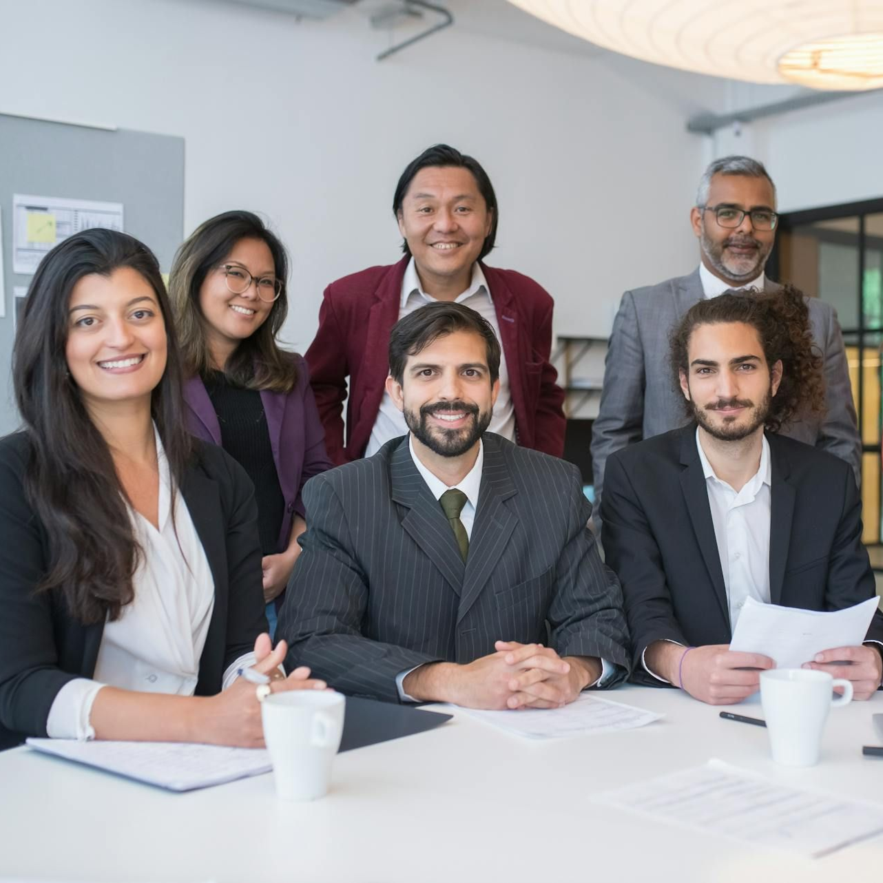 Group of six people in business attire at a table, smiling. Bright office setting, papers, and coffee cups present.