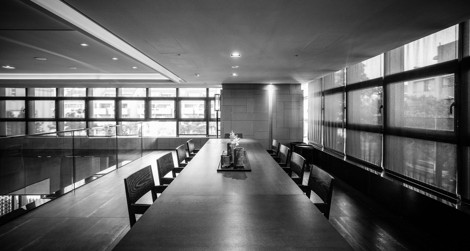 A long communal table with wooden chairs in a bright, modern office space with large windows in black and white.