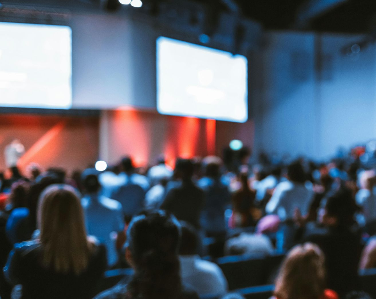 Blurred view of an audience in a large auditorium, watching a presentation on a stage with screens and red lighting.