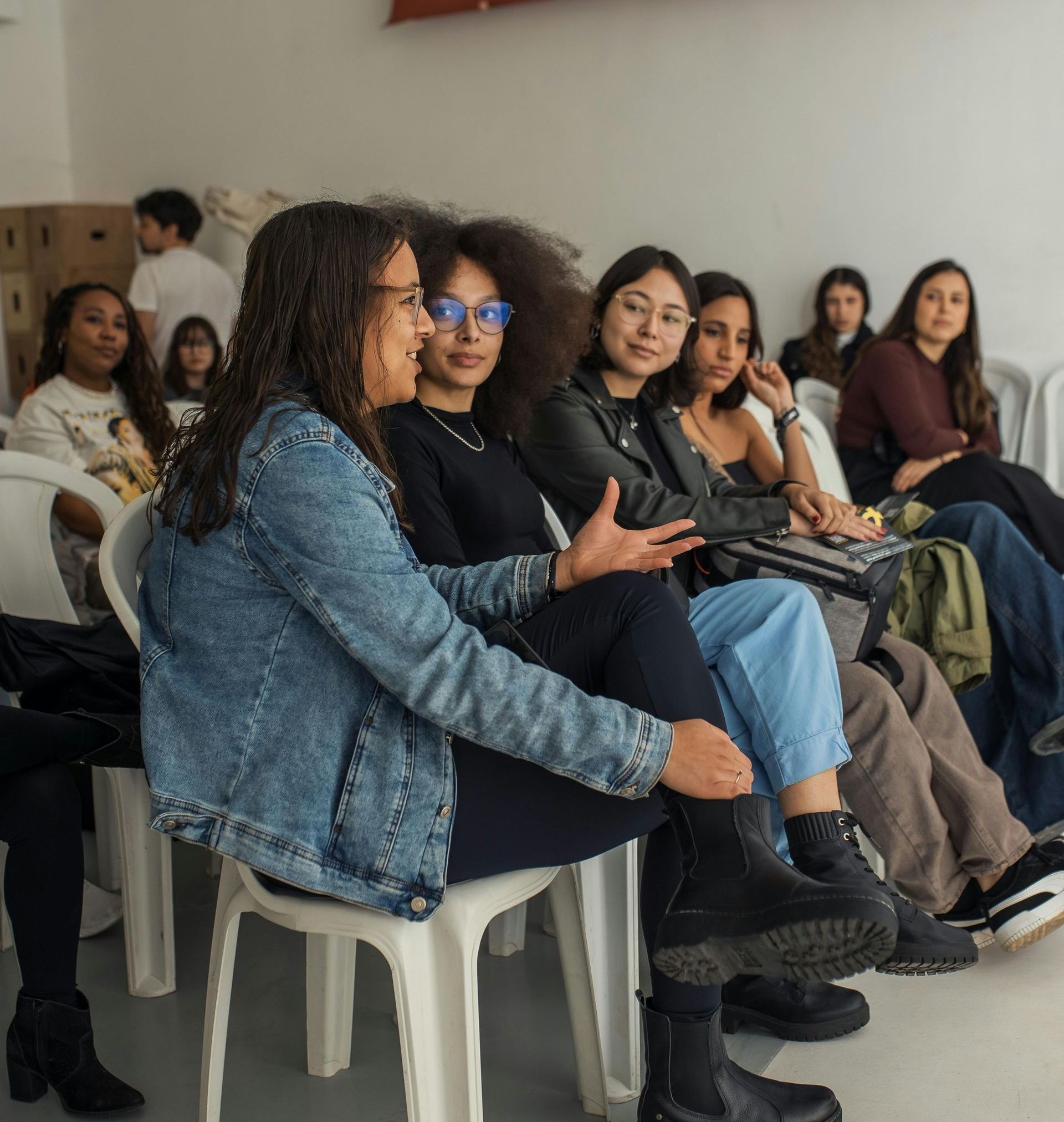 Group of women in a room, one speaking. Others listen, sitting on white chairs.