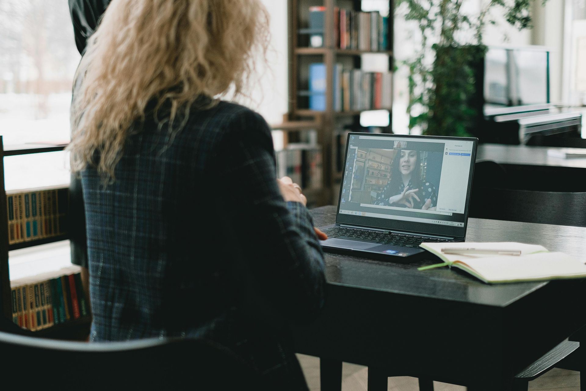 Woman at a desk with a laptop, attending a video call. Bookshelves in background.