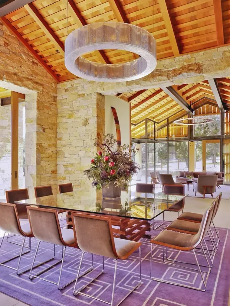 A dining room with a glass table, brown chairs, and a round pendant light, set on a purple geometric rug under wood beams.