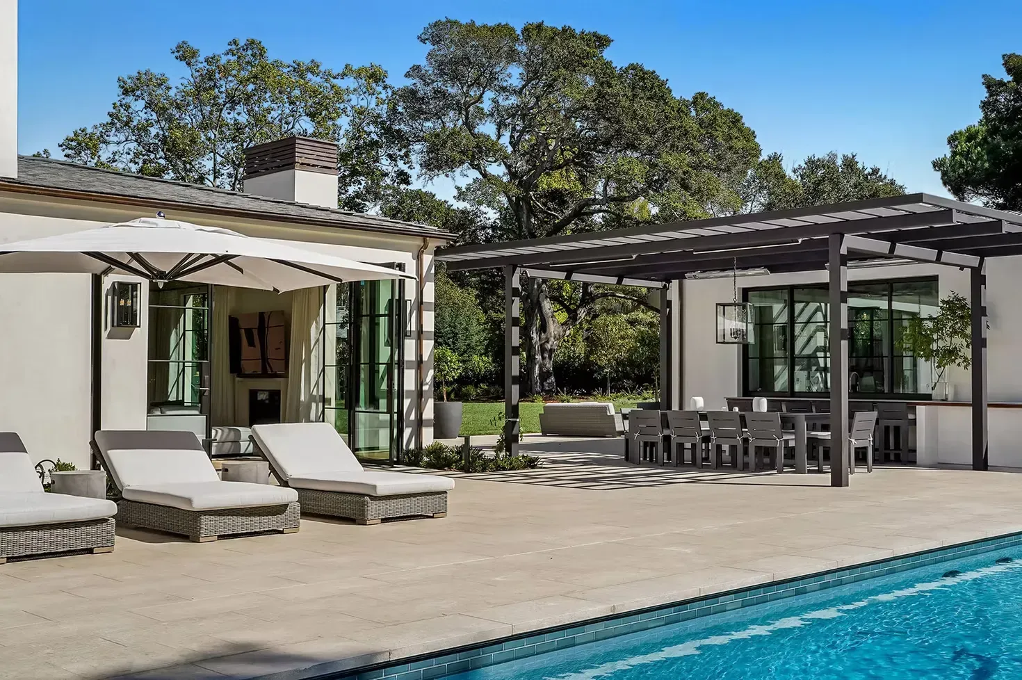 A patio beside a swimming pool features lounge chairs, an outdoor dining area under a trellis, and a white house exterior.