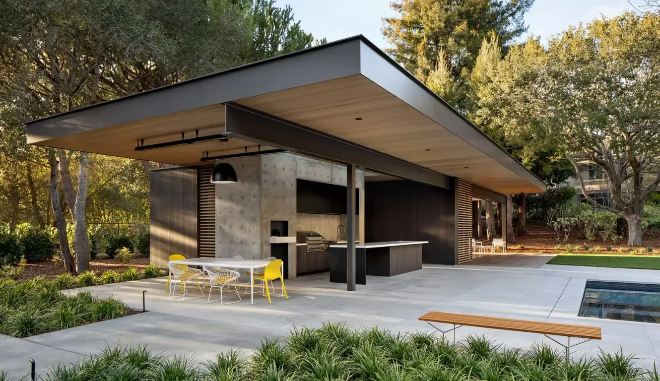 A modern, open-air pavilion with a concrete wall and kitchen, featuring a dining area beside a pool in a wooded setting.