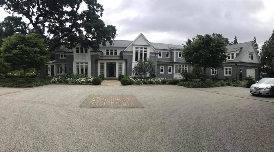 A large, two-story grey mansion with a gabled roof, white trim, and a gravel circular driveway under a cloudy sky.