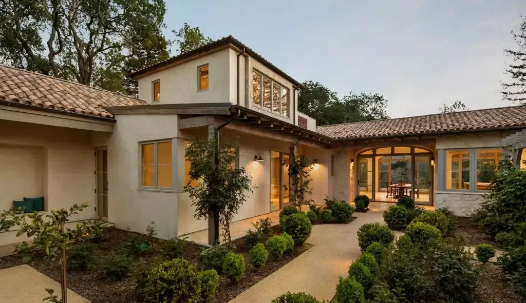 A Mediterranean-style house with a light-colored stucco exterior, tile roof, and a courtyard with manicured shrubbery.