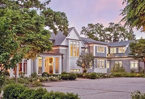 A multi-story gray shingled mansion with white trim and a gravel driveway surrounded by lush trees at dusk.
