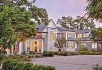A multi-story gray shingled mansion with white trim and a gravel driveway surrounded by lush trees at dusk.