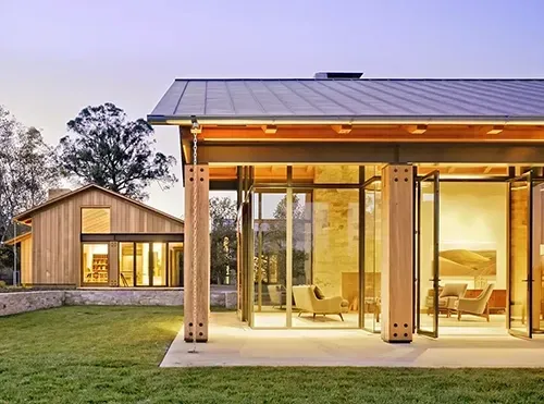 A modern house with glass walls and wood accents, illuminated from within, sits next to a smaller matching structure.