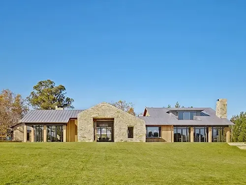 A modern, single-story stone house with a metal roof set against a clear blue sky on a grassy hill.