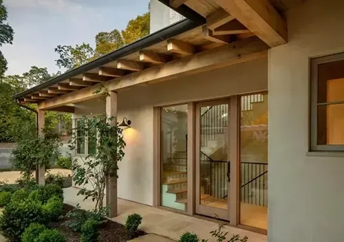 A modern, stucco house exterior featuring a wooden-beamed porch, large glass doors, and a landscaped front garden.