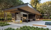 Modern outdoor kitchen with a large overhang, concrete features, dining table with yellow chairs, and pool patio.