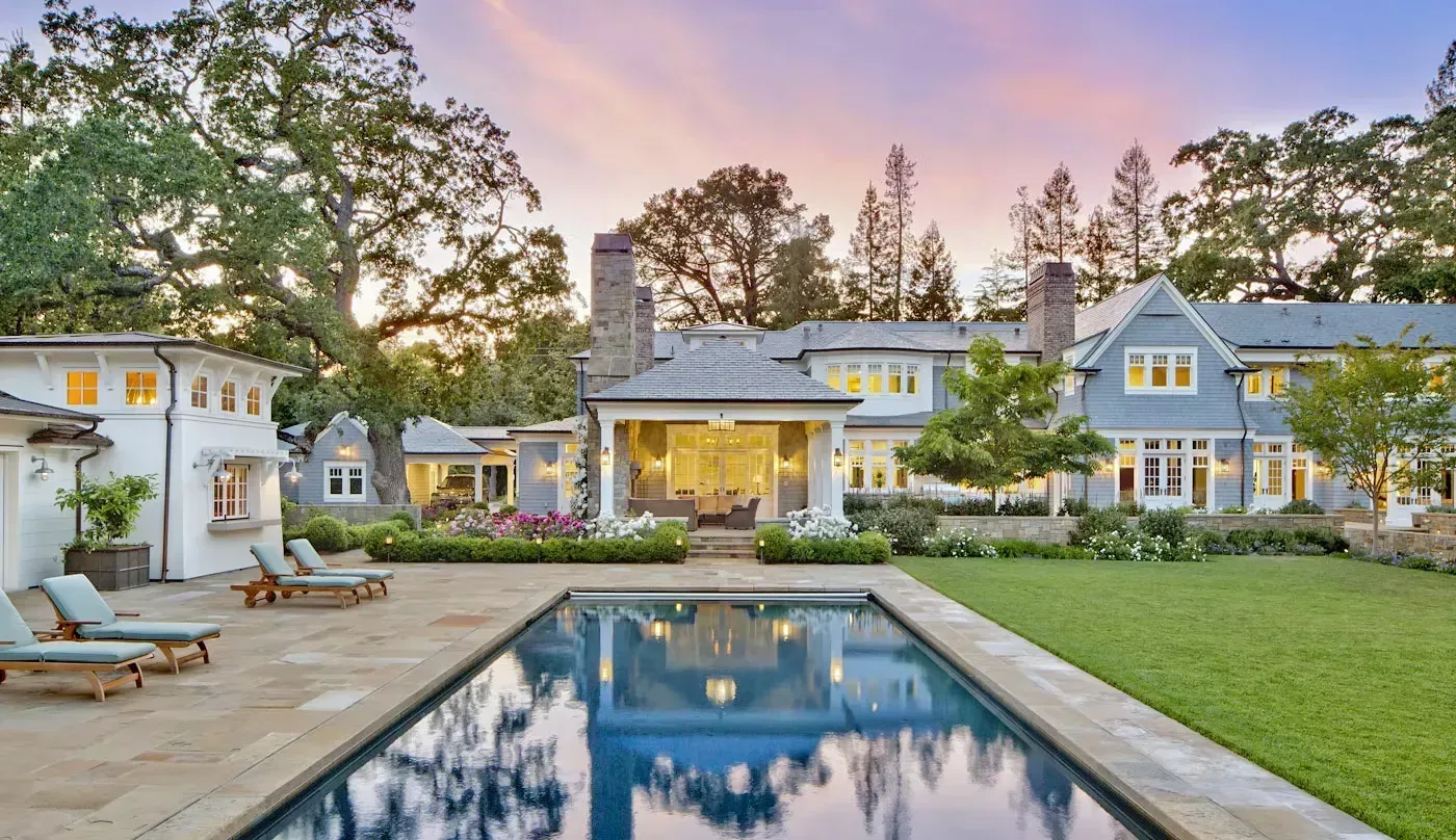 A blue shingled mansion at sunset, featuring a backyard swimming pool, stone patio, and manicured lawn.