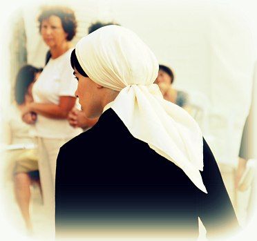 Een persoon met een witte hoofddoek en een donker gewaad, van achteren gezien, met wazige figuren op de achtergrond.