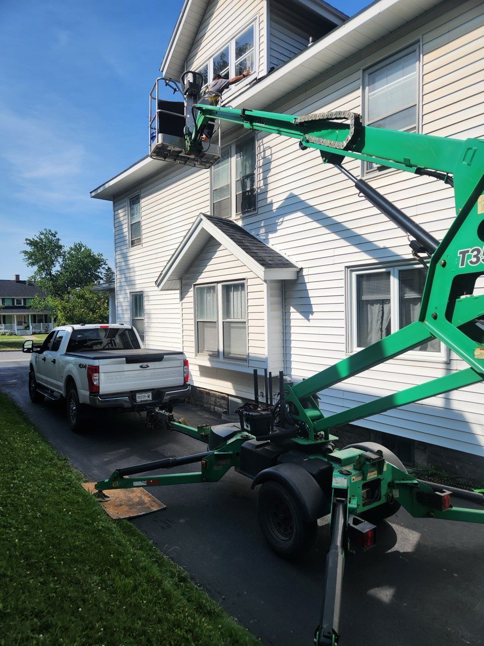 A green boom lift positioned near a white house; a person is working. A white pickup truck is parked in the driveway.