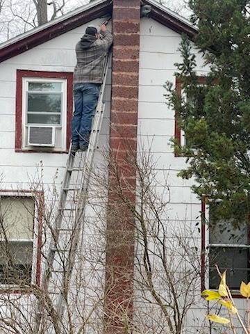 Person on a ladder, working on a chimney attached to a white house with windows and an AC unit.