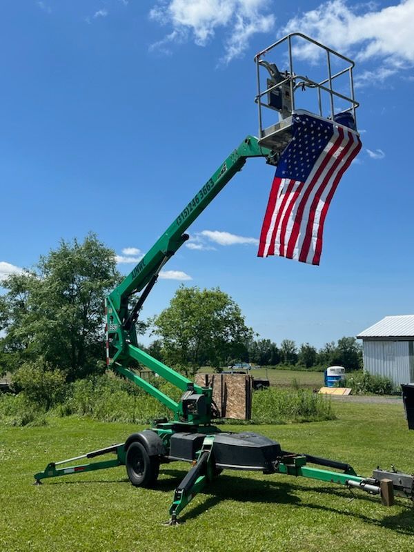 Green boom lift with American flag extended against a blue sky, in a grassy area.